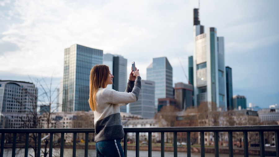 Frau macht Foto von Frankfurter Skyline auf gegenüberliegendem Ufer bei leicht bewölktem Himmel.