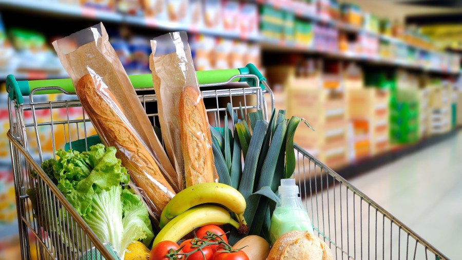 Gefüllter Einkaufswagen mit frischem Gemüse, Baguette und Bananen steht vor einem Einkaufsregal im Hintergrund.